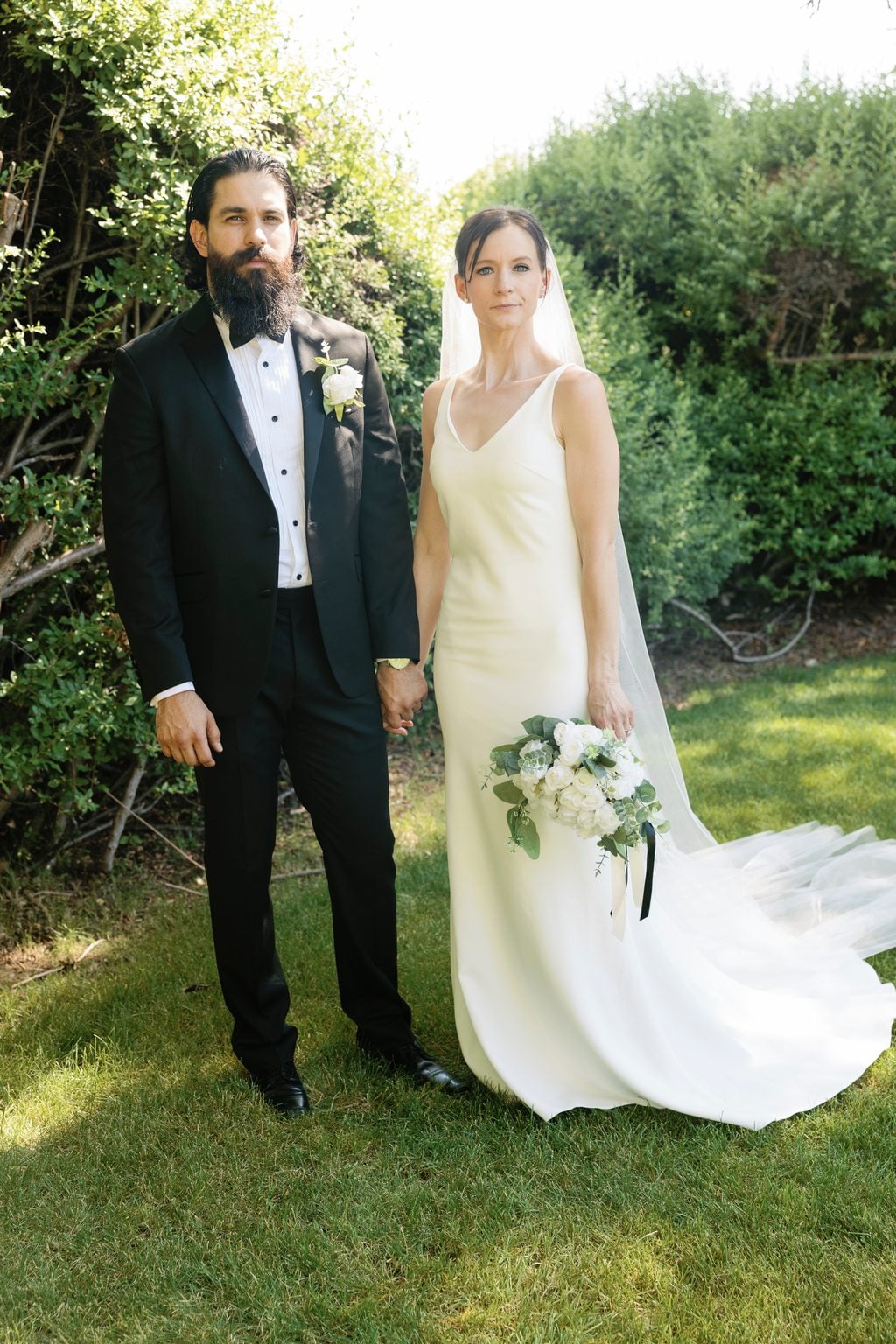 Mallory and Saint hand in hand before a garden hedge, the bride in a slip gown and long veil with a white bouquet, a black-tie wedding in Utah
