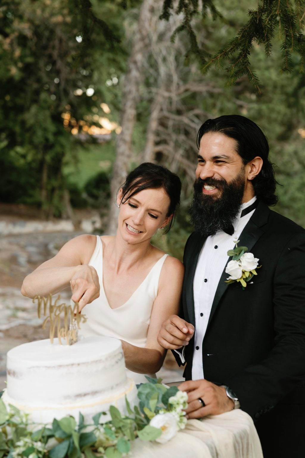 Mallory and Saint laughing as they cut the cake beneath the trees