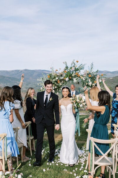 A newlywed couple walks back up a petal-strewn aisle hand in hand while guests cheer around a floral arch, with mountain views behind, at a Utah ranch wedding