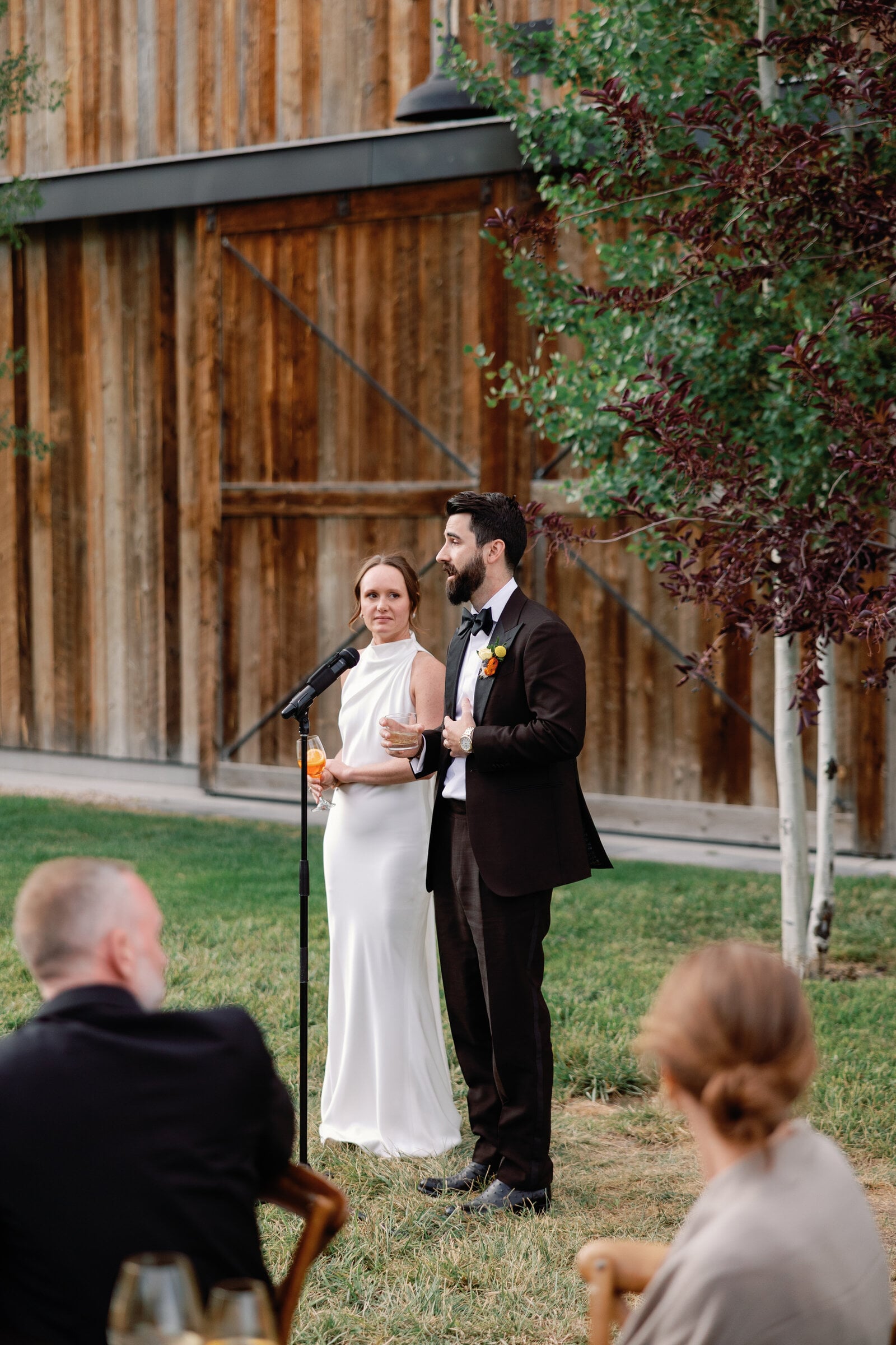 A groom speaks into a microphone with his hand on his chest while his bride stands beside him holding a drink, guests listening in the foreground, at a barn wedding in Park City, Utah