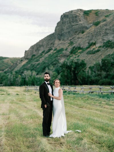 A bride and groom stand together in a meadow beneath a rock butte in soft evening light at 4U Ranch in Park City, Utah