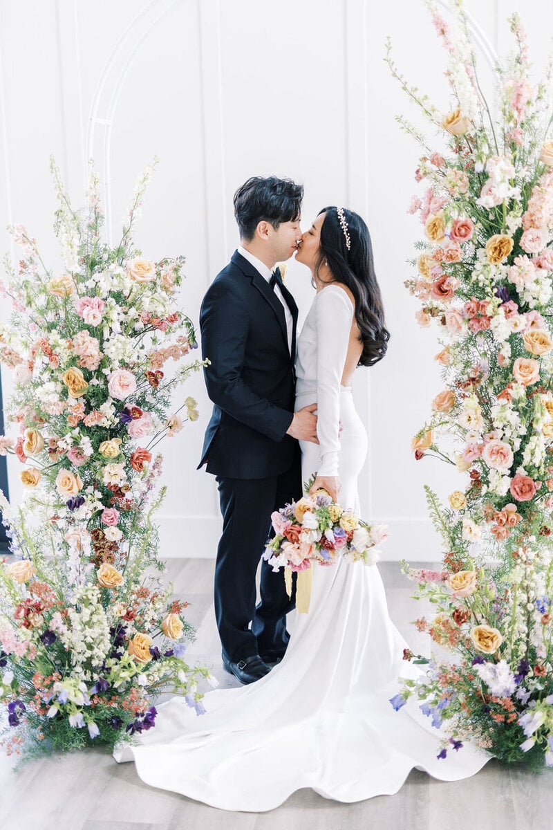 A couple kisses between two tall floral installations in muted peach, butter yellow, blush, and lavender against a white wall, the bride holding a pastel bouquet